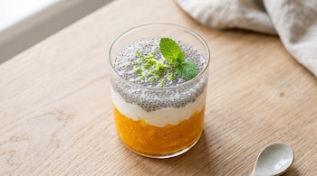 Un vaso transparente con puré de mango, yogur, semillas de chía, ralladura de lima y menta sobre una mesa de madera junto a una cuchara.