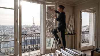 Una mujer subida a una escalera coloca papel de aluminio en el cristal de una ventana de un apartamento con vista a la ciudad y la Torre Eiffel.