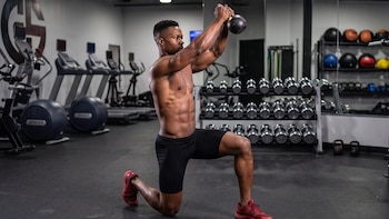 Hombre sin camiseta y en pantalones cortos negros, arrodillado sobre una pierna, levantando una pesa rusa sobre su cabeza en un gimnasio