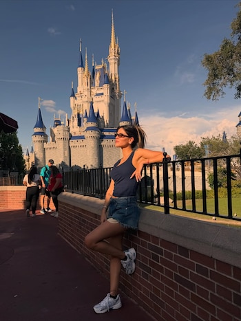 Camila Galante, con top azul y shorts de mezclilla, se apoya en una pared de ladrillo con el Castillo de Cenicienta de Disney World de fondo