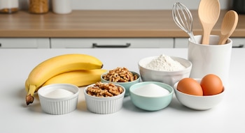 Dos bananas, tazones con nueces, harina, azúcar y dos huevos. Utensilios de cocina como un batidor y cucharas de madera en un recipiente sobre una mesada blanca.
