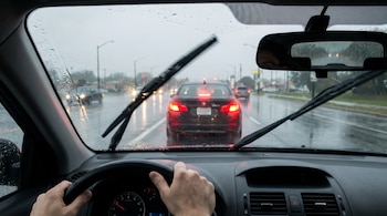 Manos en el volante de un coche bajo la lluvia. Parabrisas mojado con limpiaparabrisas. Un coche con luces de freno rojas delante en carretera mojada.
