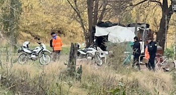 Varias personas y dos motocicletas blancas estacionadas en una zona de pastizales y árboles secos, junto a una estructura improvisada