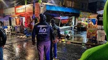 Dos personas con chamarras de la alcaldía Álvaro Obregón caminan de noche por una calle mojada frente a puestos de comida y fruterías.