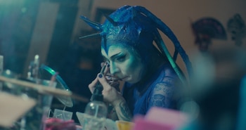 Persona con maquillaje de tono azul y tocado con púas que aplica pintura facial con un pincel frente a un espejo