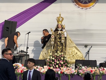 Estatua religiosa con corona y manto claro sobre un escenario con flores, rodeada por personas frente a equipos de sonido y micrófonos
