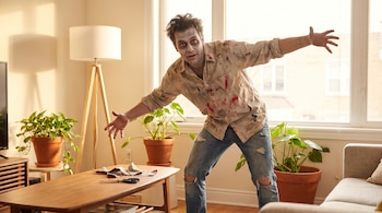 Un hombre con maquillaje de zombi, ropa rota, una mesa de centro de madera con tijeras, plantas de interior y un sofá en una sala