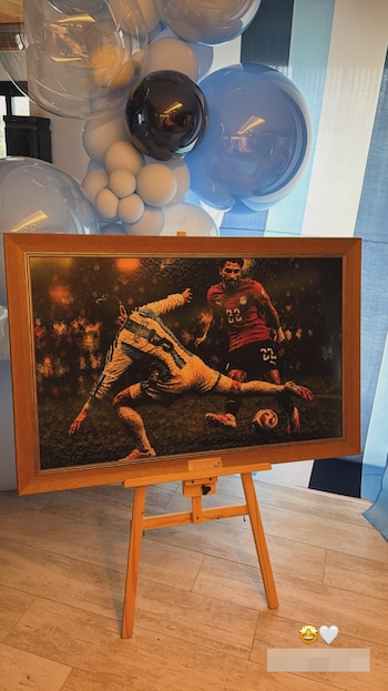 Cuadro enmarcado de un futbolista pateando un balón en un partido de fútbol, sobre un caballete de madera. Fondo con globos y telas celestes y blancas