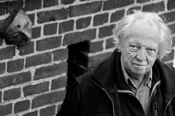 Hombre con pelo blanco, chaqueta y camisa frente a una pared de ladrillos con una pequeña escultura, fotografía en blanco y negro