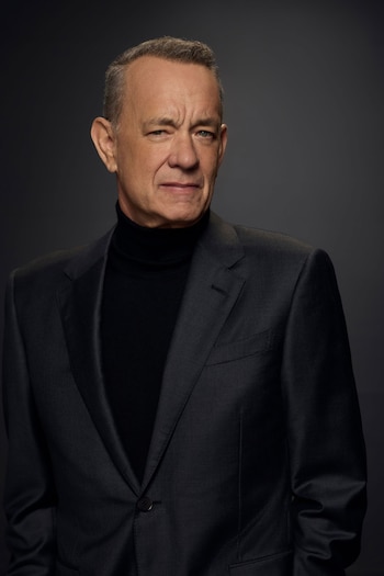 Retrato de medio cuerpo de Tom Hanks mirando directamente a la cámara, vestido con un traje oscuro y un cuello alto negro sobre un fondo gris liso
