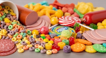 Primer plano de una mezcla de cereales coloridos, rebanadas de embutido, salchicha, caramelos de diferentes formas y galletas saladas sobre una mesa gris.