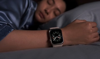 Una persona con piel trigueña duerme de lado en una cama, vistiendo una pijama azul. En su muñeca izquierda lleva un Smartwatch Huawei Luca-B19 con pantalla encendida
