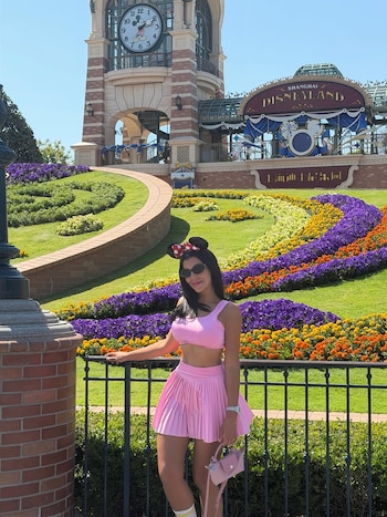 Charlotte Caniggia, con top y falda rosa, orejas de Minnie y gafas de sol, posa frente a la entrada de Disneyland Shanghai, con un reloj y jardines florales