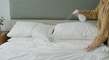 Una persona con camisa de pana beige rocía un spray blanco sobre una cama con sábanas y almohadas blancas en un dormitorio, con cabecero verde.