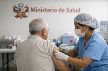 Un adulto mayor de espaldas recibe una vacuna en el brazo de una enfermera con uniforme celeste y mascarilla, frente a una pared con el logo del Ministerio de Salud de Perú.
