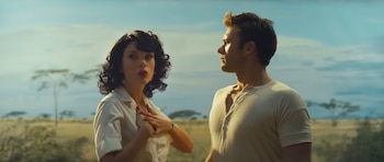 Taylor Swift, con cabello oscuro y labios rojos, y Scott Eastwood, con camiseta clara, observan a la derecha en un paisaje de sabana