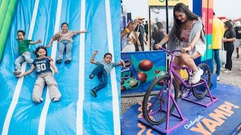 Cuatro niños en un tobogán inflable azul con franjas blancas; una mujer joven sonríe pedaleando una bicicleta estática púrpura, fondo borroso con personas