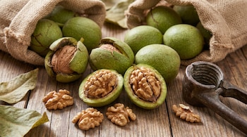 Primer plano de nueces de Castilla verdes y abiertas, frutos secos pelados, un cascanueces de metal y sacos de arpillera sobre una mesa de madera rústica.