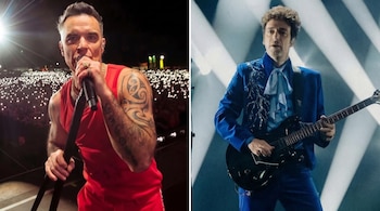 Robbie Williams con camiseta roja y micrófono en la mano junto a Gustavo Cerati con traje azul y guitarra eléctrica en el escenario.
