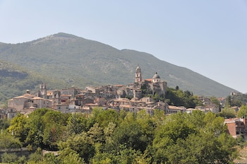 Vista de un pueblo con edificios de piedra y tejados de terracota, dominado por una iglesia con torre y cúpula, en una ladera boscosa, con una gran montaña y cielo claro al fondo