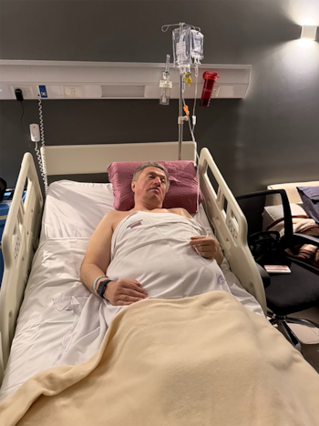 Hombre en cama de hospital con sábanas blancas y manta beige, suero intravenoso, monitor en la muñeca, almohada y una silla al lado