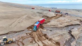 Camión de carga azul con remolque rojo transita por una trocha de tierra junto a una carretera dañada en paisaje desértico costero con maquinaria y personas