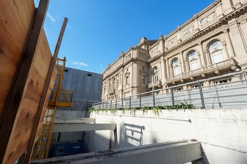 Imagen de una obra en construcción con andamios amarillos y paneles de madera en primer plano, y la fachada ornamentada del Teatro Colón bajo un cielo azul