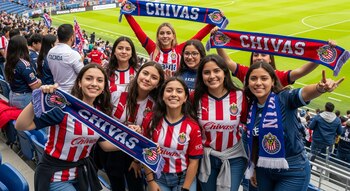 Grupo de mujeres jóvenes y adolescentes en gradas de un estadio, vestidas con camisetas de Chivas y sosteniendo bufandas del mismo equipo.