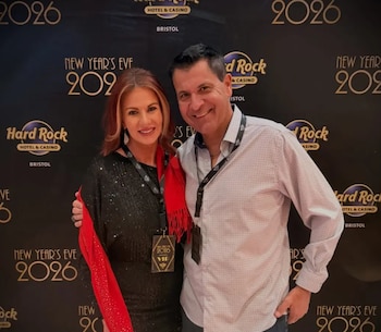 Una mujer con vestido negro y bufanda roja posa junto a un hombre con camisa clara frente a un fondo con logotipos