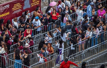 Decenas de personas caminan y hacen fila entre vallas metálicas de seguridad frente a un cartel publicitario rojo