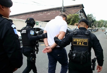 Agentes policiales con chalecos tácticos escoltan a un hombre esposado con camiseta blanca y pantalón oscuro en una calle