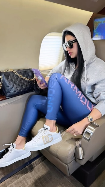 Una mujer sentada en un asiento de avión con un hoodie gris, leggings azules, zapatillas blancas, gafas de sol y un teléfono morado. Hay un bolso negro al lado