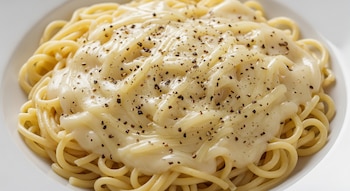 Fideos amarillos con salsa blanca cremosa y pimienta negra molida espolvoreada, servidos en un plato blanco hondo sobre un fondo claro.