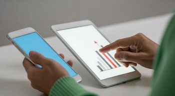 Primer plano de manos de piel asiática sosteniendo un smartphone y una tablet. La tablet muestra una gráfica de barras con flecha roja de crecimiento, el smartphone tiene pantalla azul.