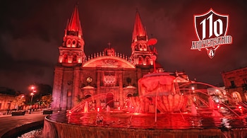 Fachada de una catedral gótica iluminada en rojo intenso, con dos torres y una fuente en primer plano que lanza chorros de agua rojiza. Escudo del 110 aniversario.