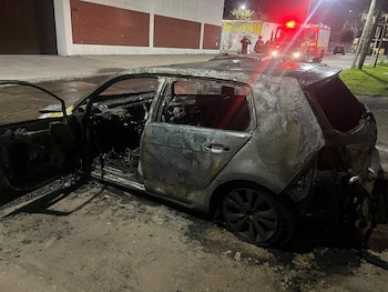 Un automóvil calcinado con la puerta delantera abierta en una calle nocturna, con un camión de bomberos al fondo
