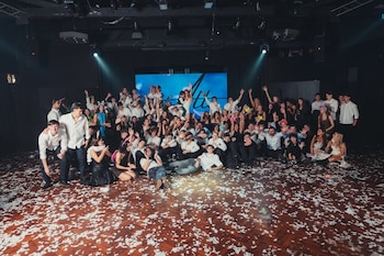 Gran grupo de jóvenes y adultos posando en una pista de baile con papel picado y una pantalla azul de fondo durante una fiesta