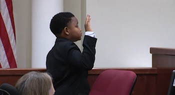 Un niño afroamericano con traje negro levanta la mano derecha en un tribunal junto a una bandera de Estados Unidos
