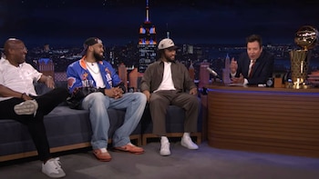 Jimmy Fallon en un escritorio con un trofeo dorado de baloncesto, frente a tres hombres en un sofá. Fondo de horizonte urbano nocturno con edificio iluminado