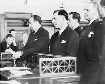 Hombres en trajes formales en un tribunal; uno firma documentos en una mesa, mientras otros observan de pie. Fotografía en blanco y negro mostrando la audiencia