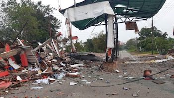 Ruinas de una estructura con escombros rojos y blancos en primer plano, con un techo metálico verde y blanco parcialmente colapsado y una carretera con desechos al fondo