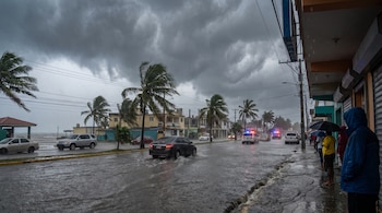 Calle inundada con vehículos transitando, palmeras al viento, personas con paraguas en la acera y cielo nublado por tormenta.