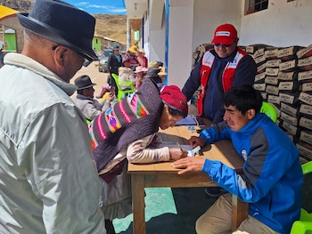 Un hombre de espaldas, una mujer con manta y gorro rojo firmando en una mesa, un joven, y un hombre con gorra y chaleco de Cofopri. Montañas y sacos al fondo