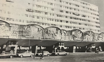 Edificio alto en blanco y negro con una fachada inferior adornada con grandes relieves artísticos, vehículos antiguos estacionados al frente y algunas personas caminando