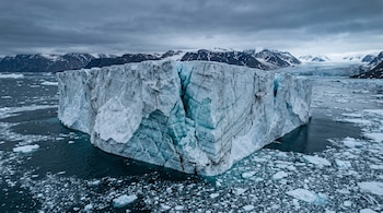 Bloque masivo de hielo con una grieta vertical flota rodeado de pequeños fragmentos sobre el mar cerca de montañas.