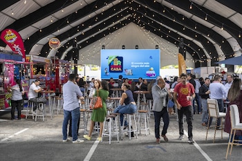 Interior de carpa con techo a rayas y luces. Personas de pie y sentadas en mesas altas con taburetes. Pantalla al fondo con logo 'Expo CASA'. Camiones de comida a la izquierda