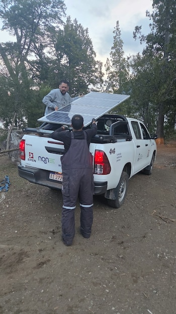 Dos hombres acomodan un panel solar sobre la caja de una camioneta blanca rodeada de árboles