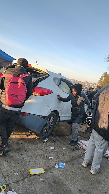 Automóvil blanco dañado en la parte trasera con rueda elevada sobre una roca, personas alrededor, mochila roja, suelo sucio y cielo azul