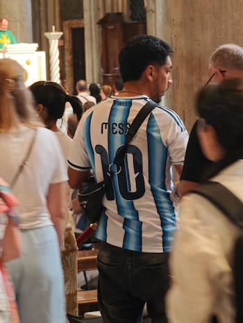 Hombre de espaldas con camiseta de fútbol de Argentina, número 10, nombre Messi. Dentro de un edificio religioso con gente y un sacerdote de vestimenta verde