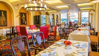 Comedor elegante con mesas cubiertas de blanco, sillas de madera y rojas, candelabros, cuadros de estilo antiguo en las paredes y ventanales con vista exterior